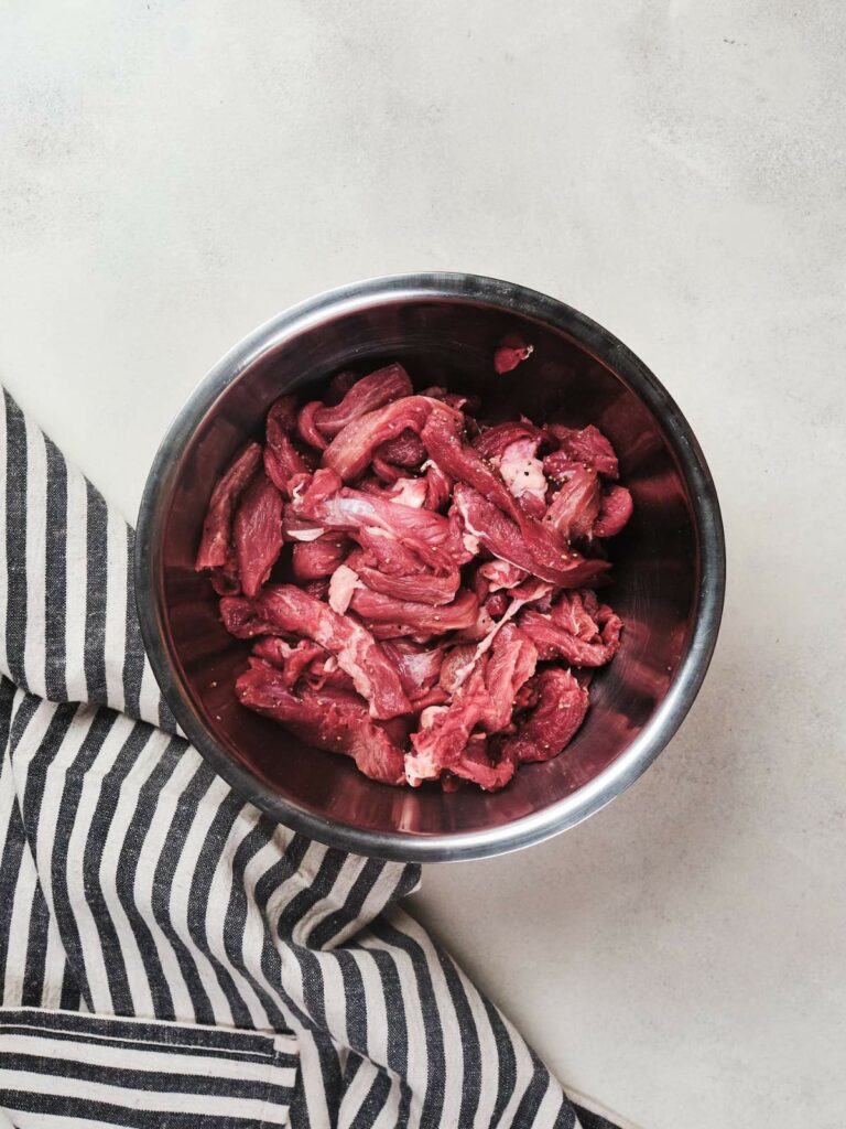 Raw sliced beef and broccoli in a stainless steel bowl on a striped cloth and concrete background.