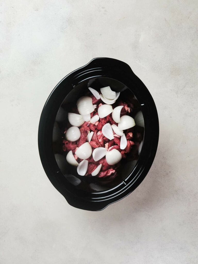 A slow cooker filled with raw beef chunks and broccoli florets, viewed from above on a light grey surface.