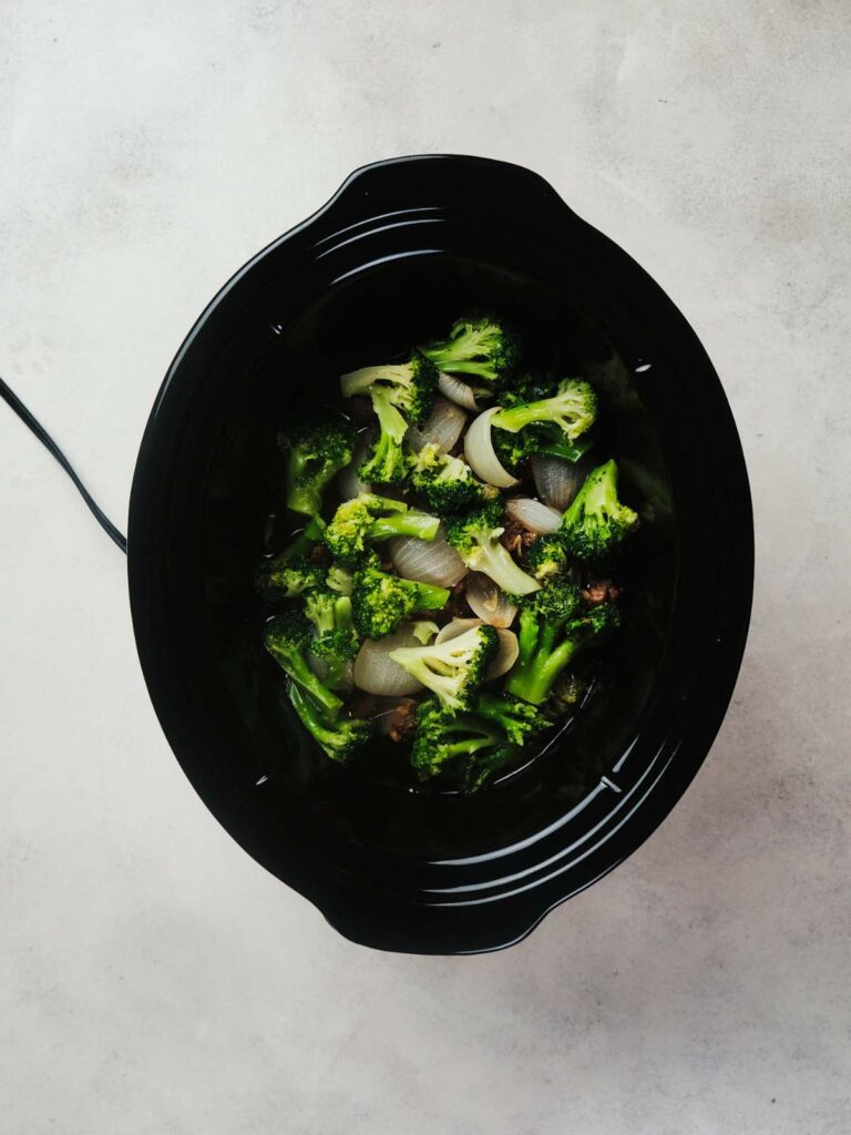 A black crockpot containing a healthy keto beef and broccoli dish on a light gray surface.