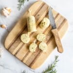 Slices from a roll of rosemary garlic butter on wooden board with a knife.