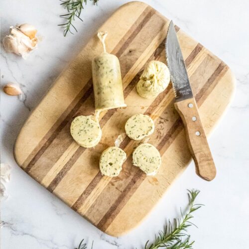 Slices from a roll of rosemary garlic butter on wooden board with a knife.