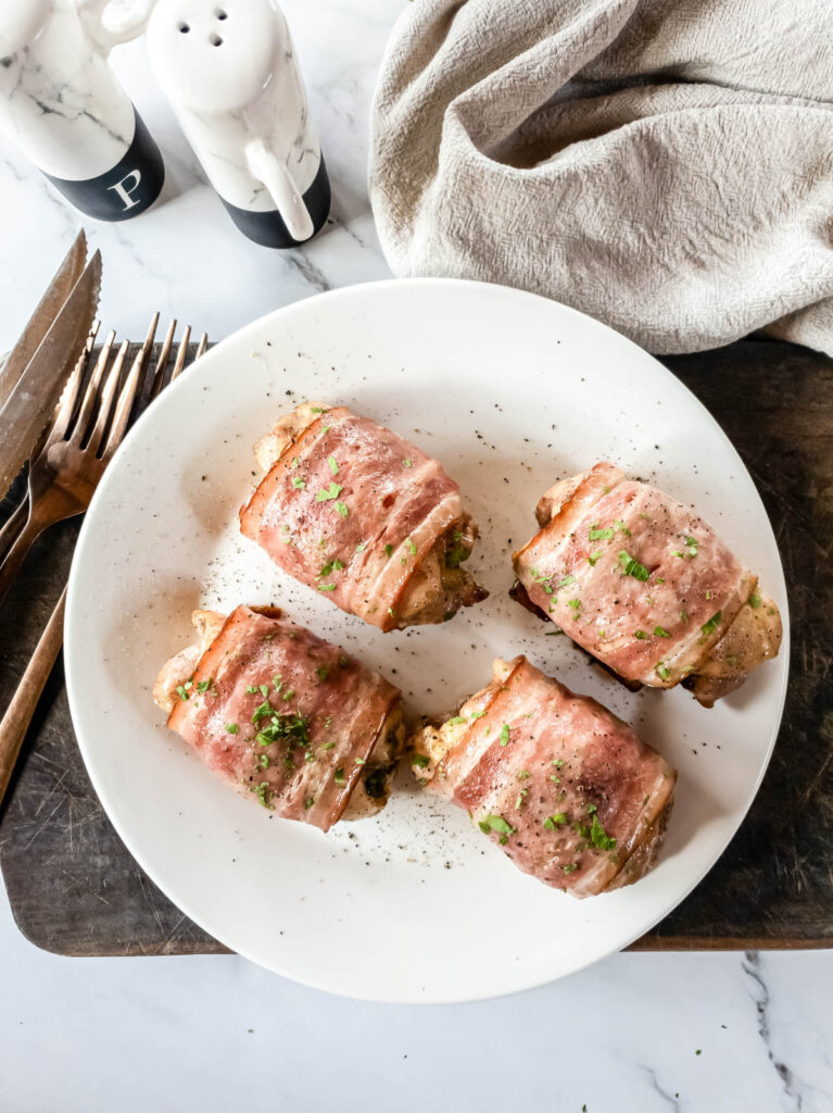 Four bacon wrapped chicken thighs on plate on wooden board.