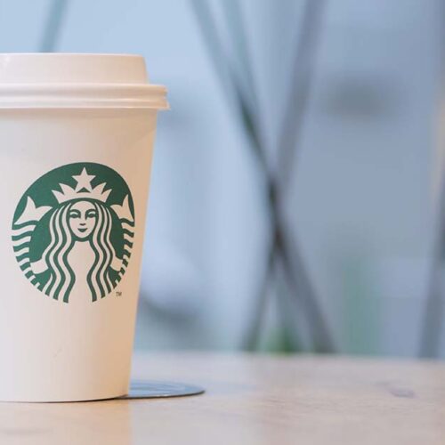 A sugar-free Starbucks coffee cup sits on a table, lights blurred in background.