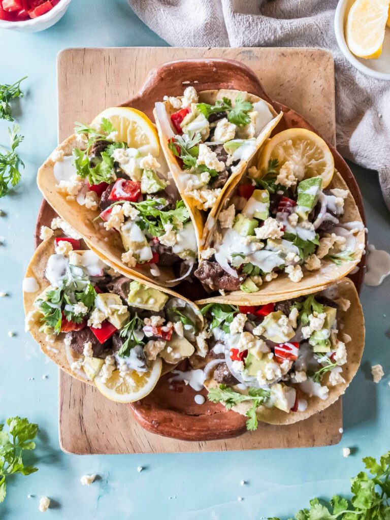 Five beef heart tacos on a wooden cutting board with fresh ingredients.