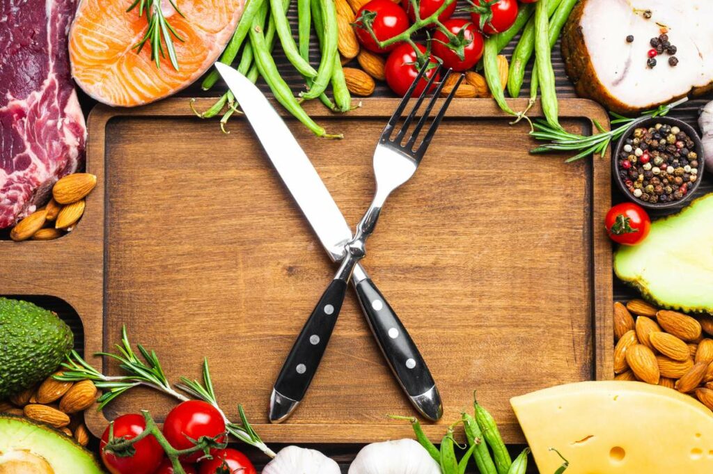 A knife and fork crossed over each other on a cutting board with freshingredients around the sides.