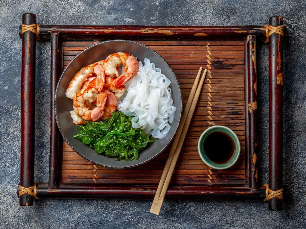 A bowl of shrimp and vegetables with Shirataki noodles on a wooden tray with chopsticks.