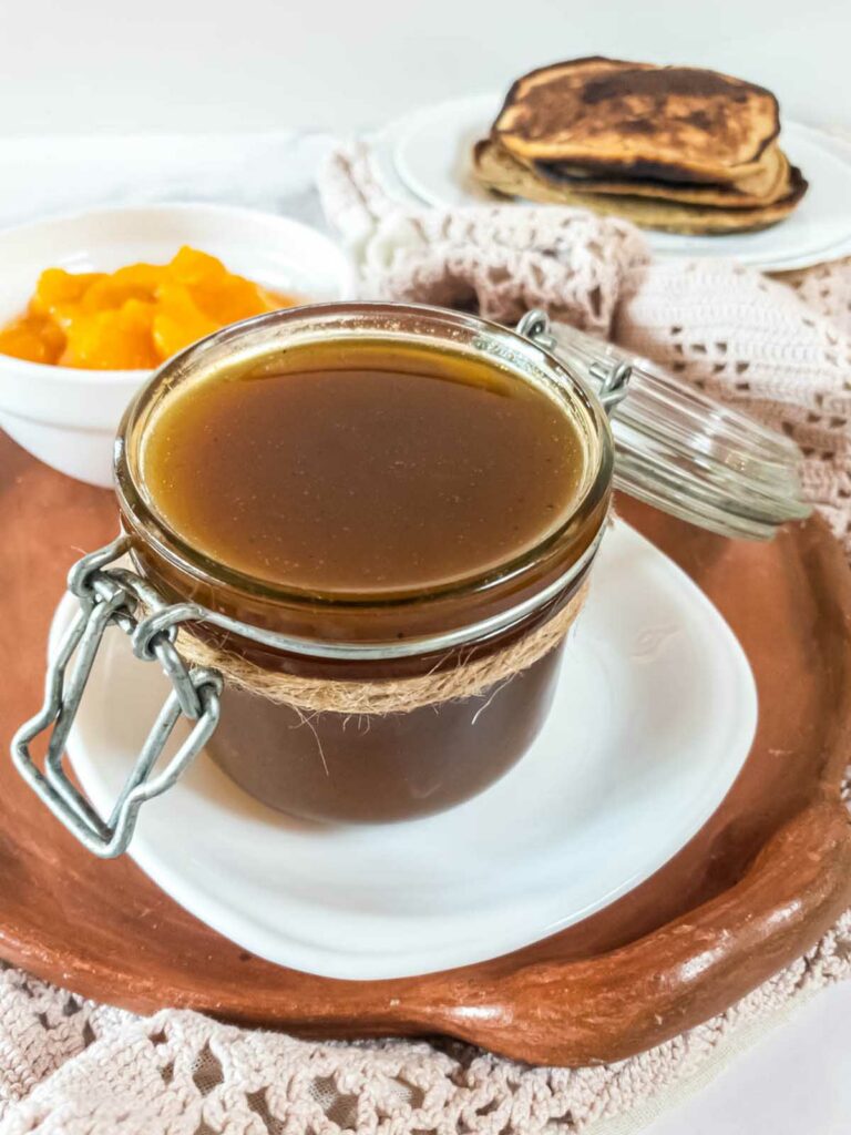 A jar of pumpkin syrup next to a plate of pancakes.