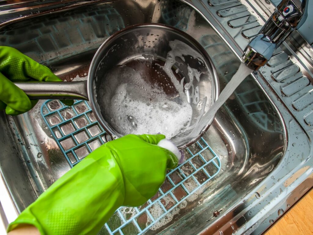 A person in green gloves washing a steel pan in a kitchen sink.