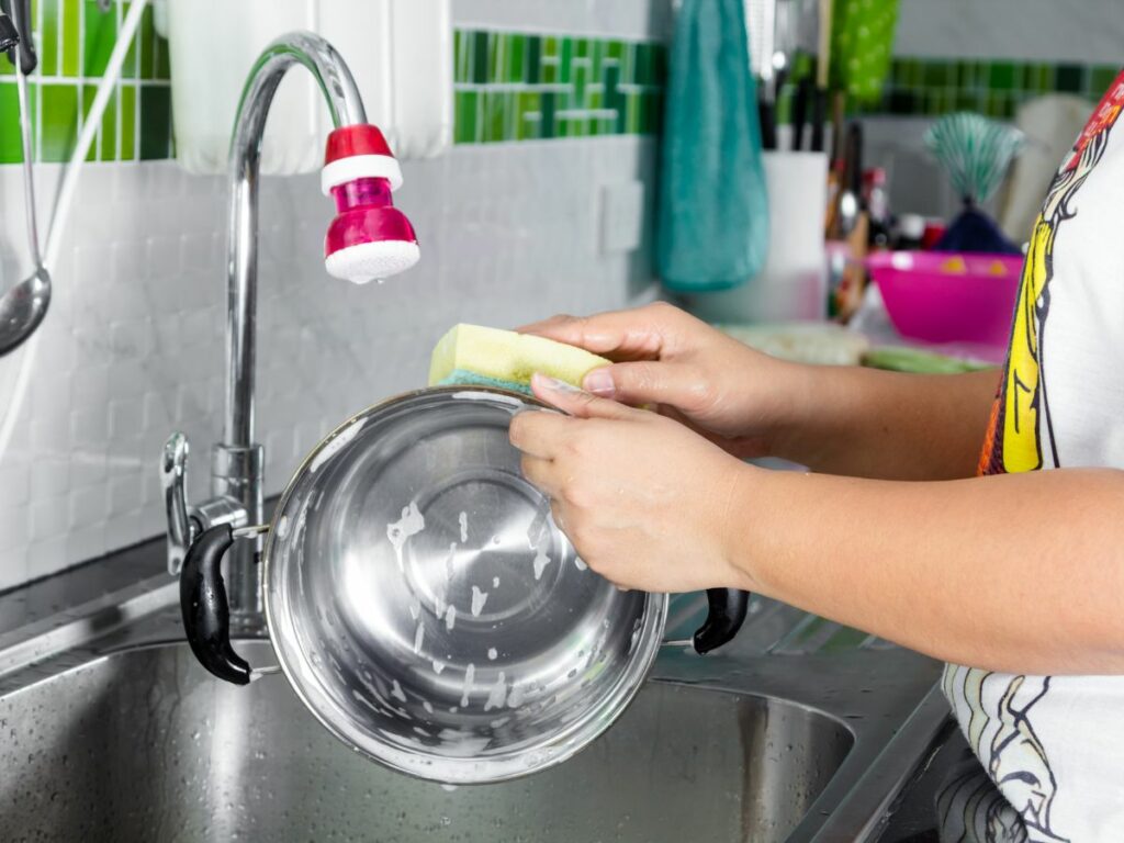 A woman is cleaning a stainless steel pan in a kitchen sink.
