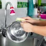 A woman is cleaning a stainless steel pan in a kitchen sink.