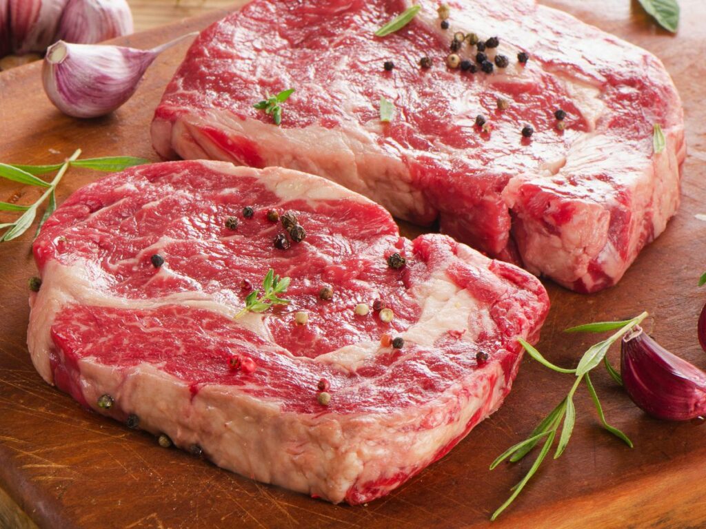 Two ribeye steaks on a cutting board with herbs and garlic.