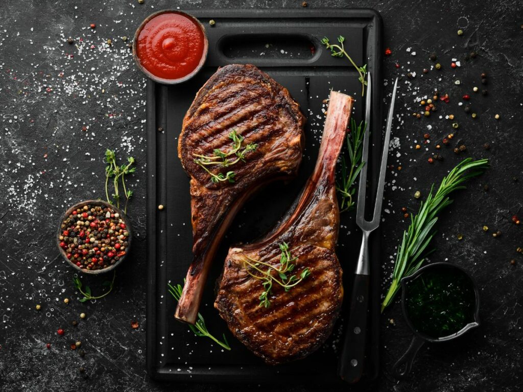 Two grilled ribeye steaks on a cutting board with herbs and spices.