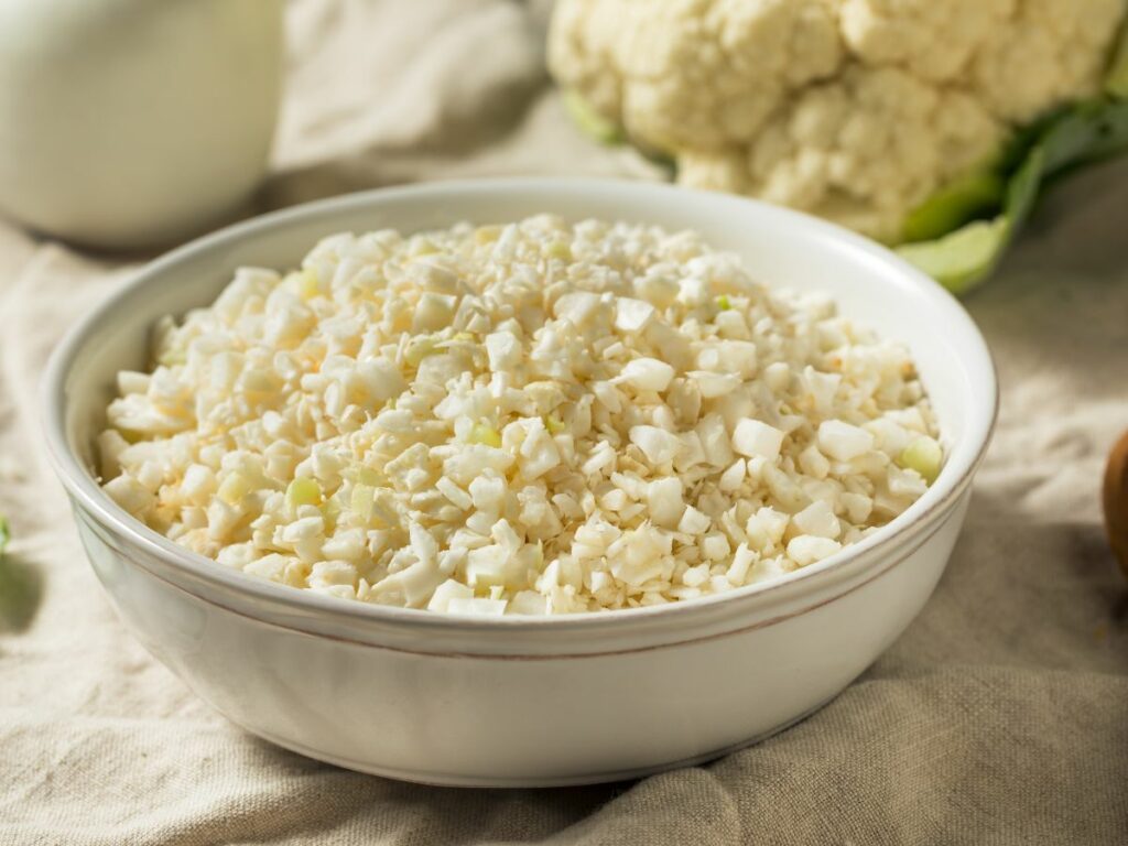 Cauliflower rice in a bowl next to a fresh cauliflower.