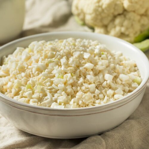 Cauliflower rice in a bowl next to a fresh cauliflower.