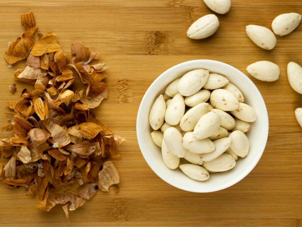 A bowl of white almonds and a bowl of shelled almonds on a wooden table.