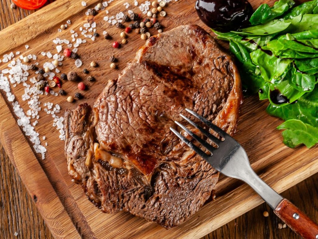 A rib steak on a wooden cutting board with vegetables and a fork.