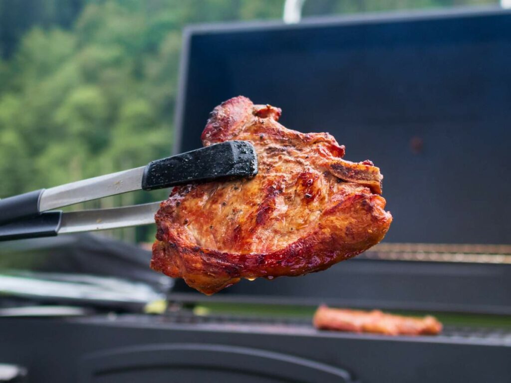 A person is holding a piece of meat with tongs over a grill.