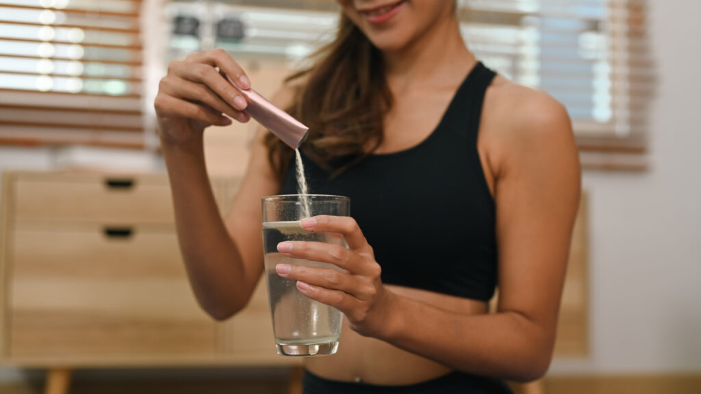 A woman in workout attire pouring a collagen powder supplement into a glass of water.