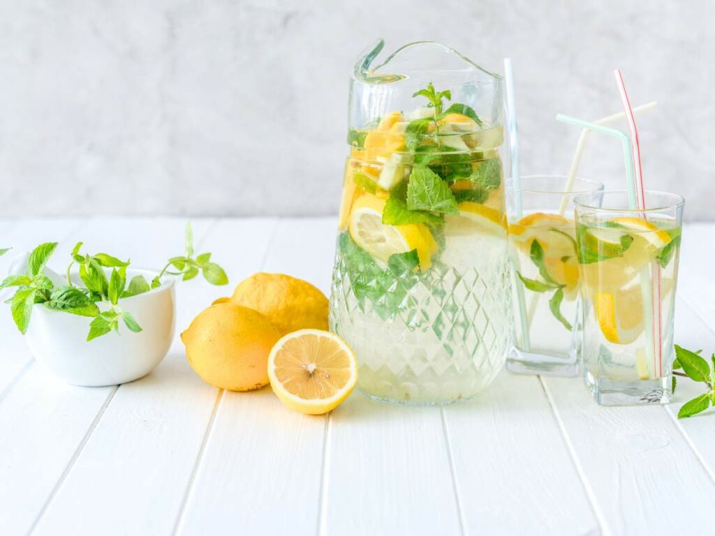 A pitcher and a glass of lemon-infused water garnished with mint leaves, accompanied by fresh lemons and mint on a white wooden surface.
