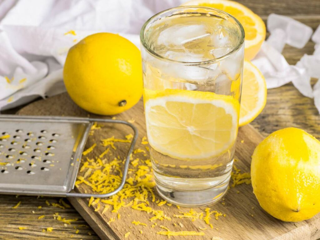 A glass of water with lemon slices, surrounded by whole lemons, lemon zest, and a zester on a wooden surface.