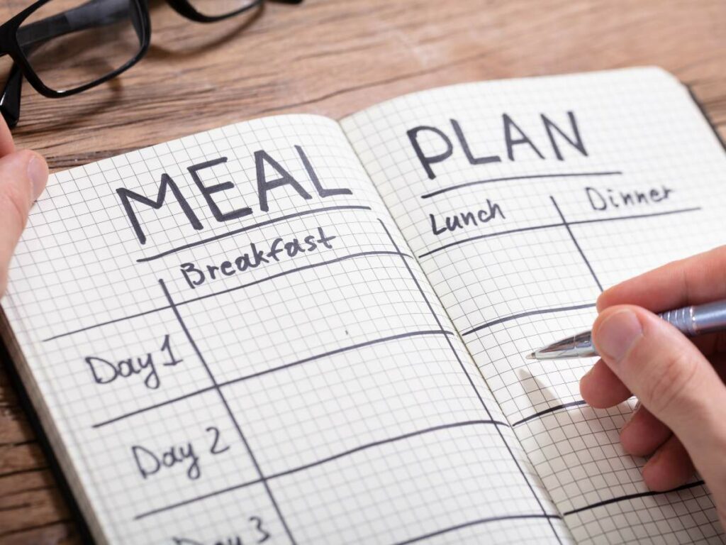 A person writes in a notebook labeled "meal plan" with sections for breakfast, lunch, and dinner.
