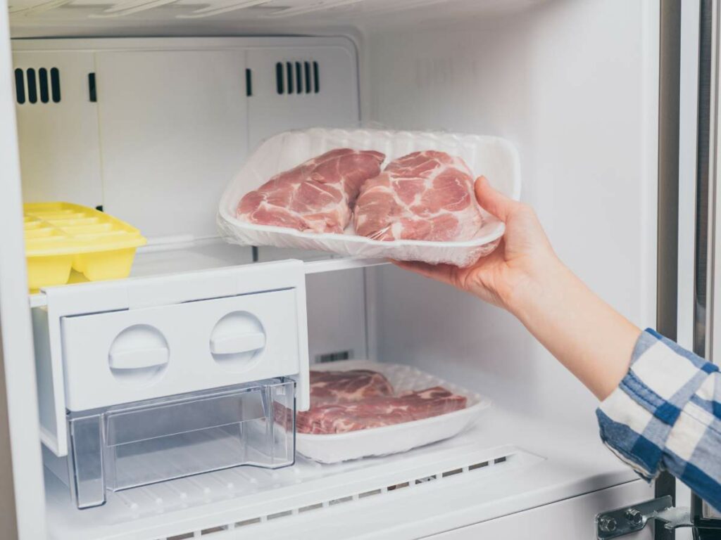 A person placing a tray of raw pork chops into a refrigerator.