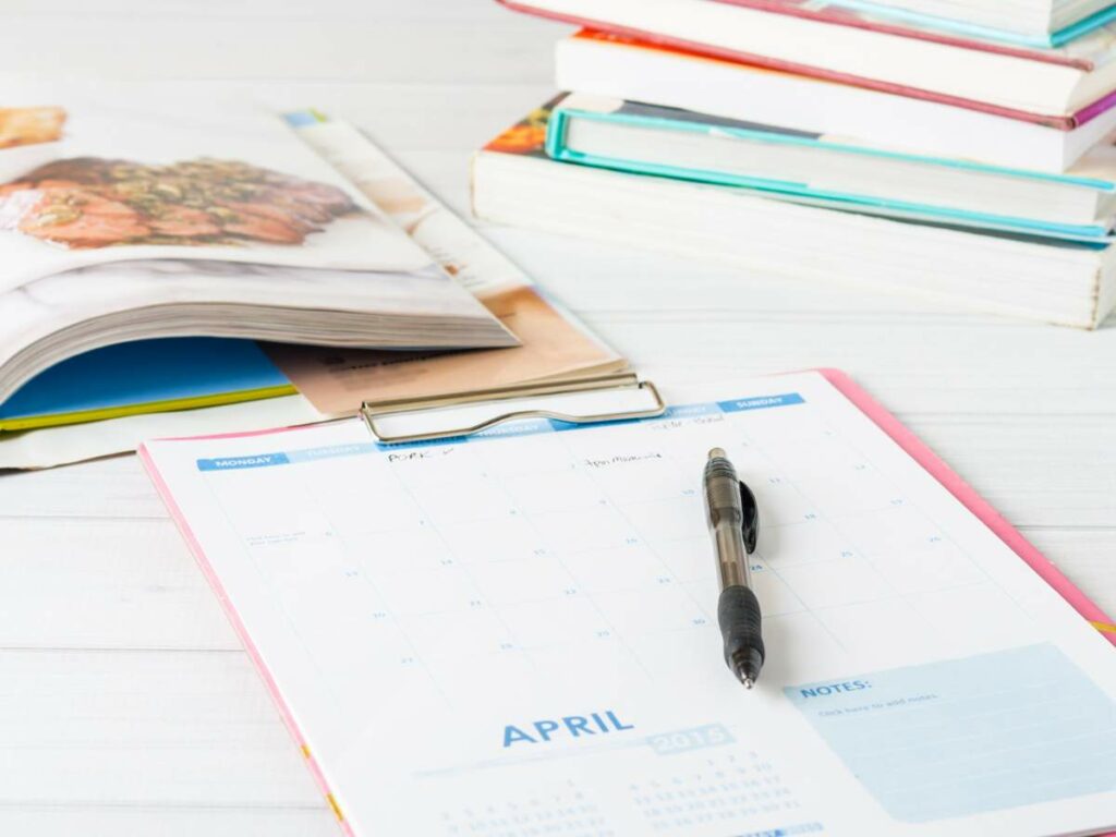 A monthly planner for april rests on a white table with a pen on top, next to a stack of books and an open magazine.