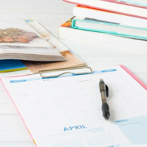 A monthly planner for april rests on a white table with a pen on top, next to a stack of books and an open magazine.