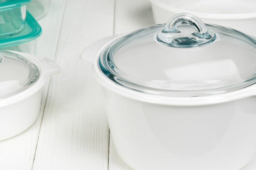 Various sizes of white ceramic cookware with glass lids on a wooden surface.