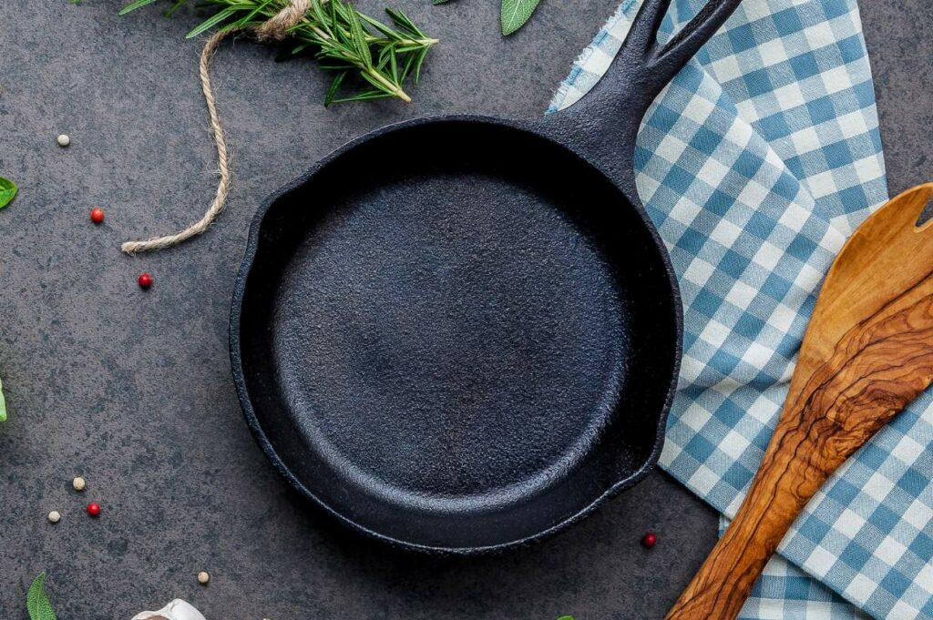 Empty cast iron skillet on a dark countertop with herbs and spices scattered around and a wooden spatula to the side.