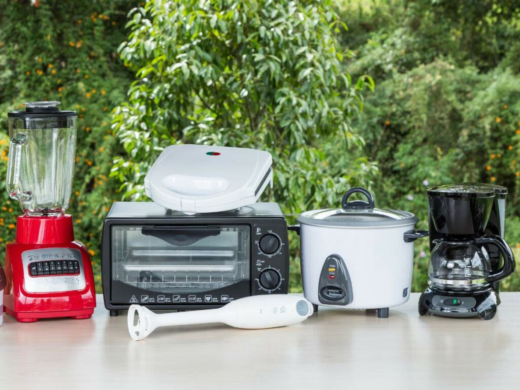 A collection of small kitchen appliances on a table, including a blender, toaster, sandwich maker, toaster oven, slow cooker, hand blender, and coffee maker, with green foliage in the background.