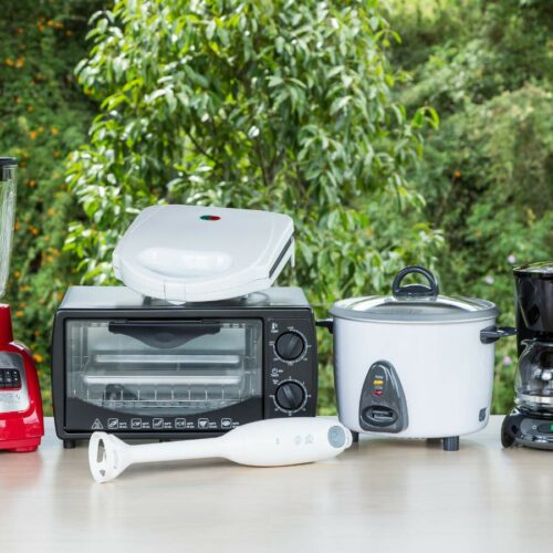 A collection of small kitchen appliances on a table, including a blender, toaster, sandwich maker, toaster oven, slow cooker, hand blender, and coffee maker, with green foliage in the background.