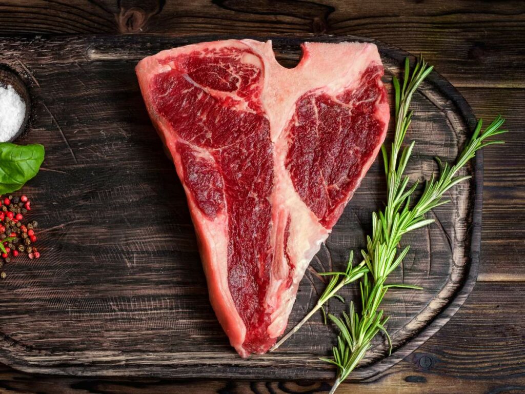 A t-bone steak on a wooden cutting board with herbs and spices.