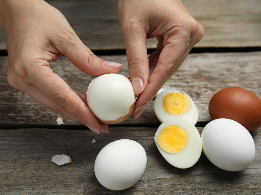 Person peeling a hard-boiled egg with other eggs in various states of peeling on a wooden surface.