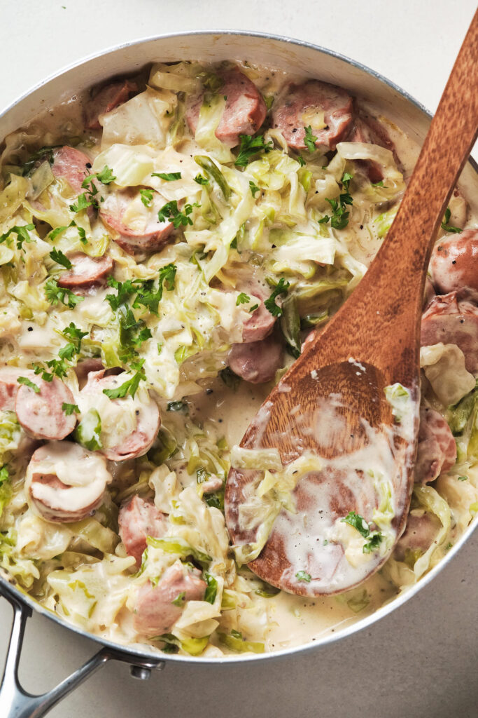 Sautéed cabbage and sliced sausages in a creamy sauce, garnished with chopped parsley in a skillet with a wooden spoon.