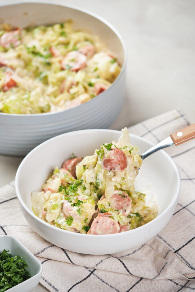 A bowl of sliced sausages and cabbage in Alfredo sauce, next to a skillet.