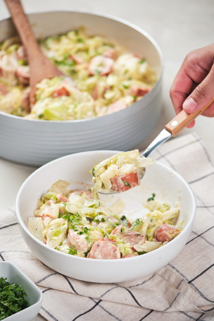 A person serving cabbage and sausage Alfredo from a pot to a white bowl.