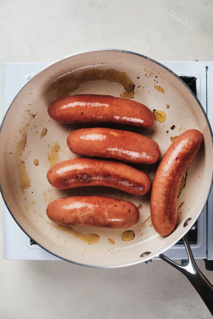 Five sausages cooking in a white frying pan, set on a stove top.