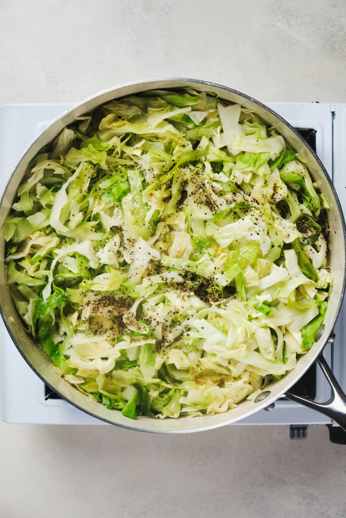 A pan of shredded cabbage seasoned with herbs and pepper, cooking on a stove.