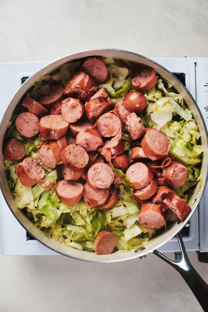 Sautéed sliced sausages and chopped cabbage in a skillet on a cooktop.