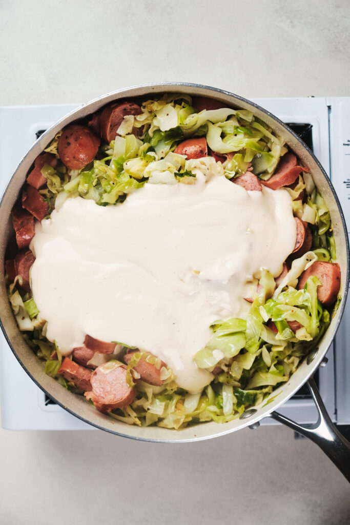 A skillet of sliced sausages and chopped cabbage topped with a Alfredo sauce.