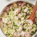 Skillet with creamy cabbage and sliced sausage dish, garnished with parsley, wooden spoon inside.