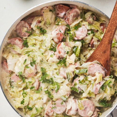 Skillet with creamy cabbage and sliced sausage dish, garnished with parsley, wooden spoon inside.