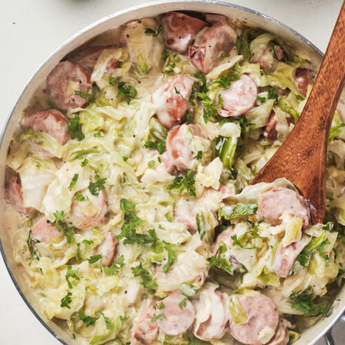 Skillet with cabbage and sausage alfredo, garnished with parsley, wooden spoon inside.