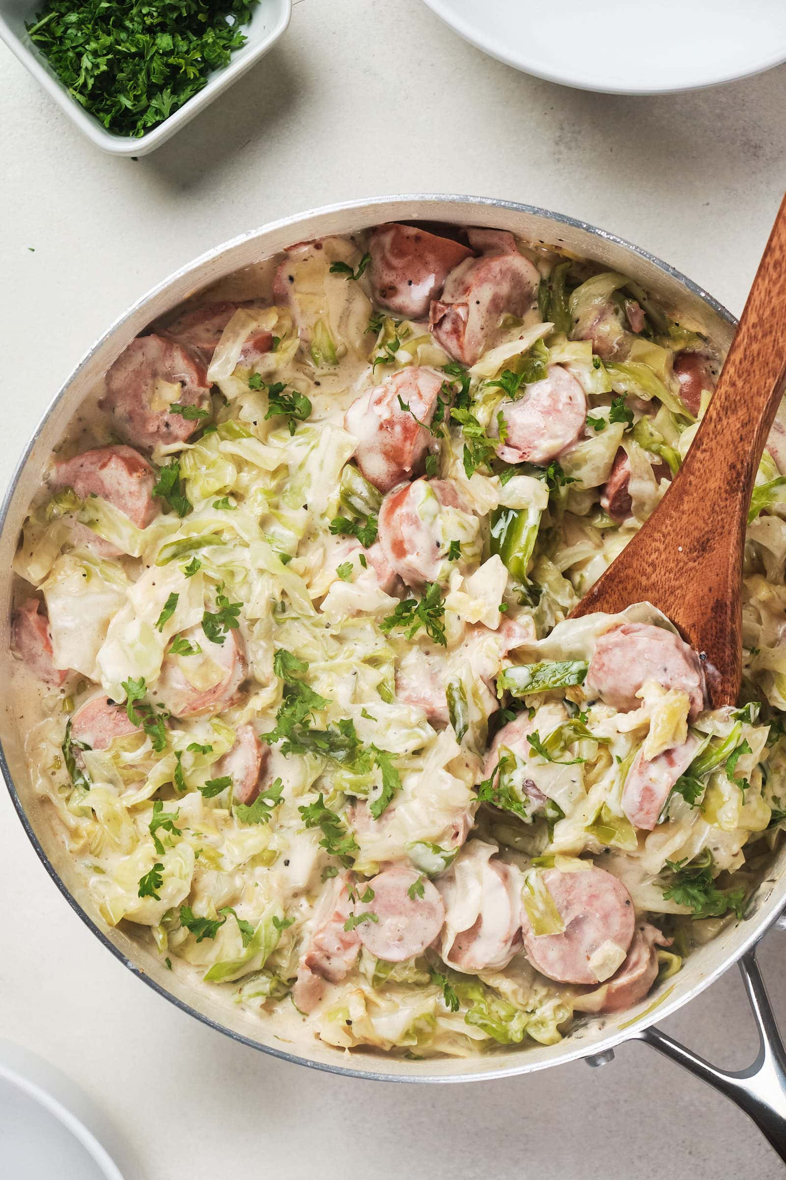 Skillet with creamy cabbage and sliced sausage dish, garnished with parsley, wooden spoon inside.