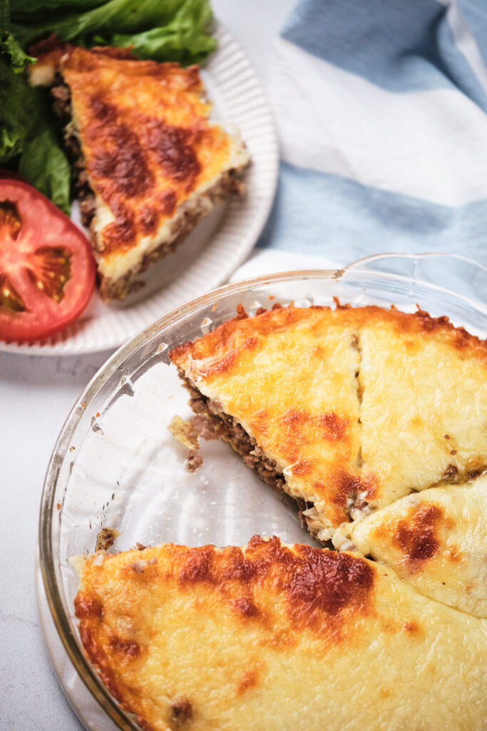 A glass pie dish containing slices of cheeseburger pie, with one slice served on a plate.