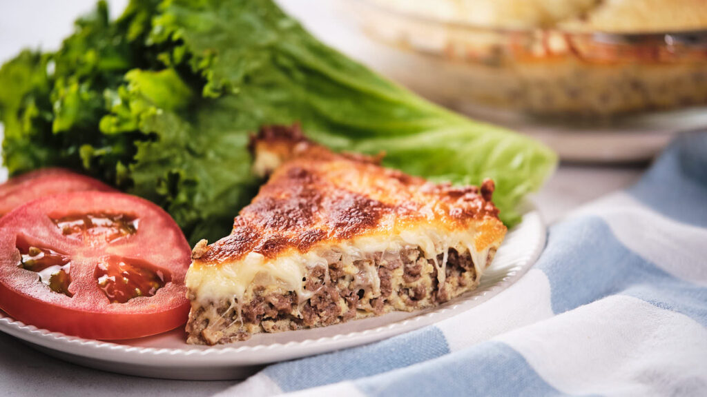 A slice of cheeseburger pie, accompanied by fresh tomato slices and lettuce on a white plate.