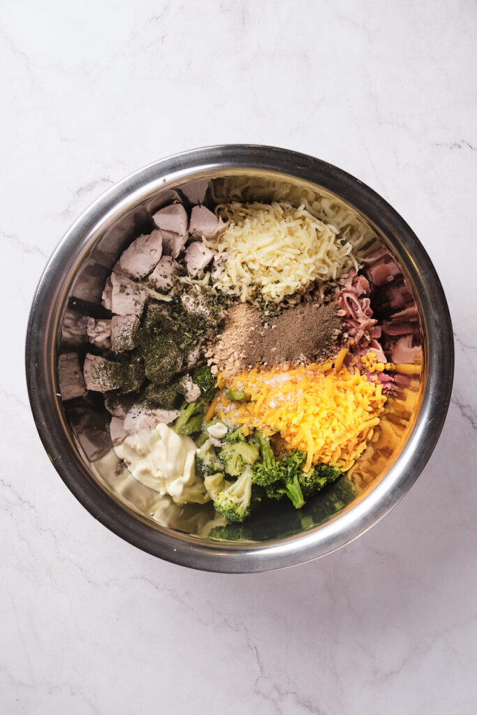 A bowl containing assorted ingredients like chicken, cheeses, vegetables, and spices on a marble surface.