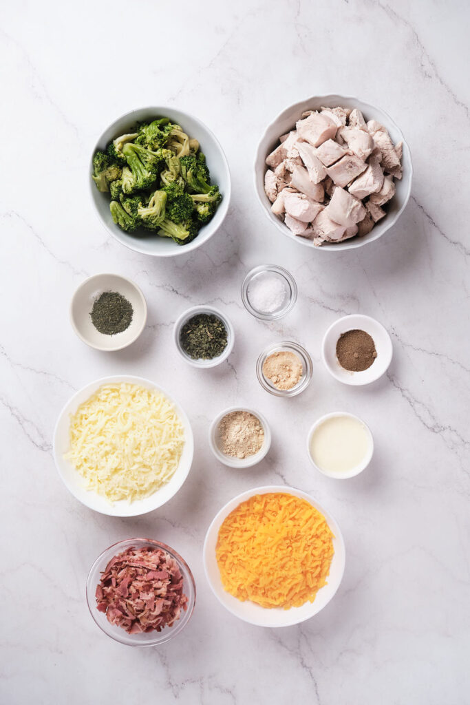 Ingredients including bowls of broccoli, cooked chicken, cheese, and spices.