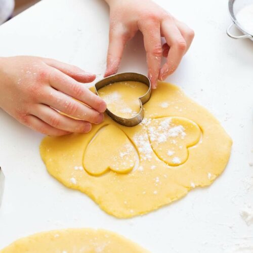 Hands pressing a heart-shaped cookie cutter into rolled dough, with flour scattered around.